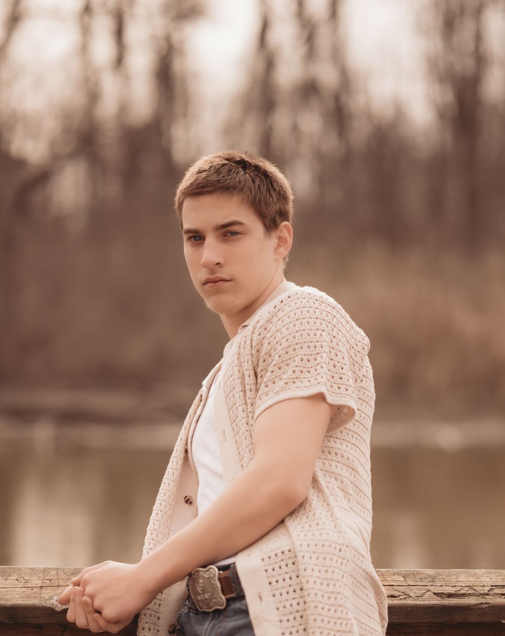 A person in a white patterned shirt leans against a railing near a lake with trees in the brown tinted background.