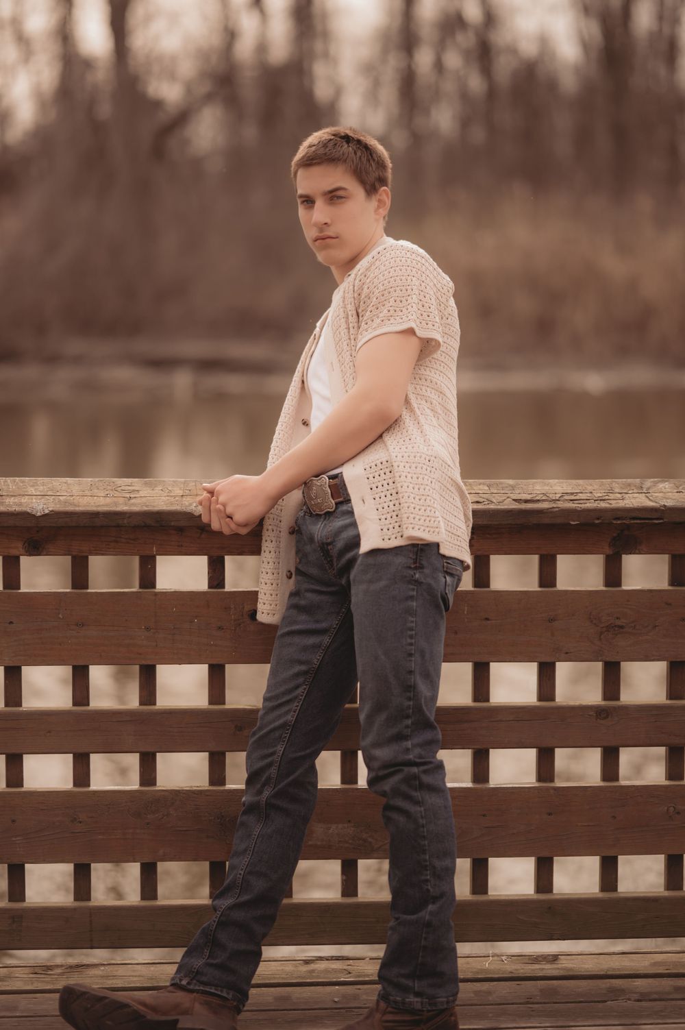 Person in cream knit shirt and dark jeans poses on wooden bridge deck during sunset.
