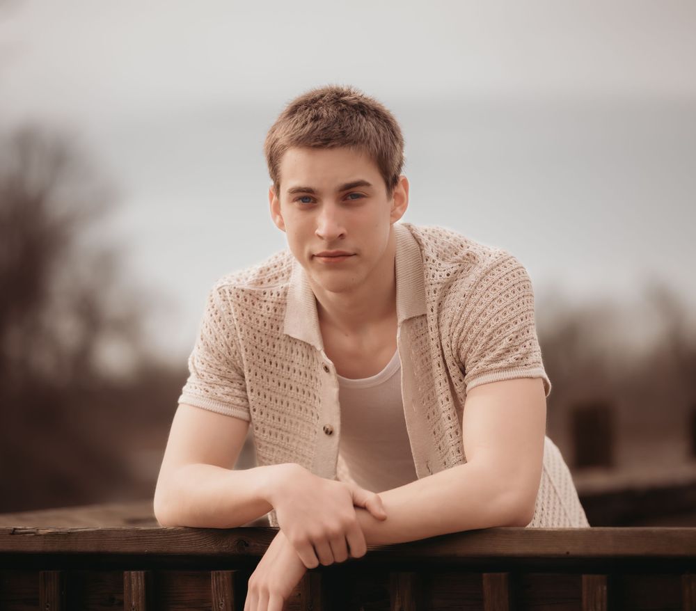 A person in a beige knit cardigan leans against a wooden railing in a moody outdoor setting.