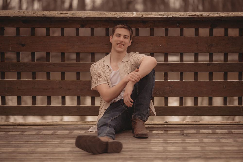 A person in casual clothing sits relaxed on a wooden bench during a sepia-toned outdoor photoshoot.