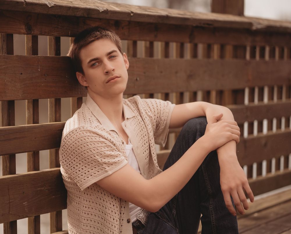 A young person in a beige knit sweater sits casually against a wooden slatted fence in a portrait-style photo.