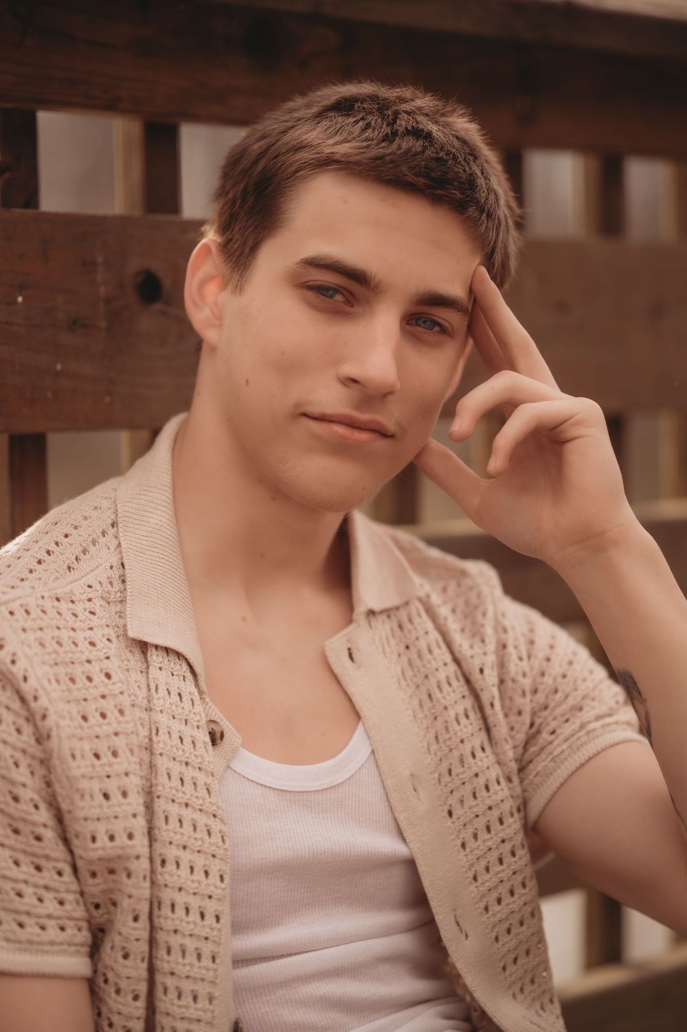 Person wearing beige knit cardigan and white tank top poses with thoughtful expression against wooden backdrop.