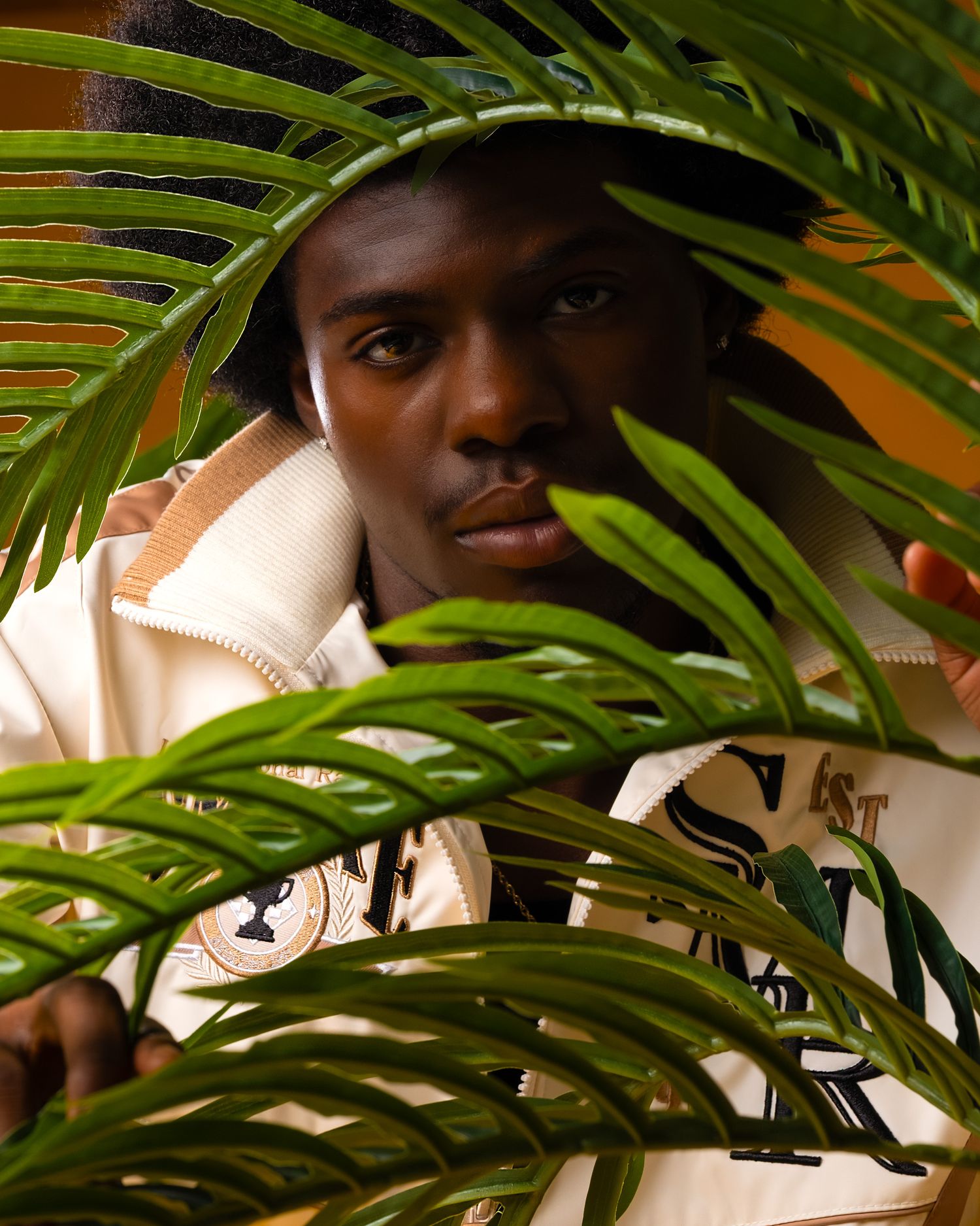 Artistic fashion portrait through tropical palm fronds with warm lighting and a cream-colored jacket.