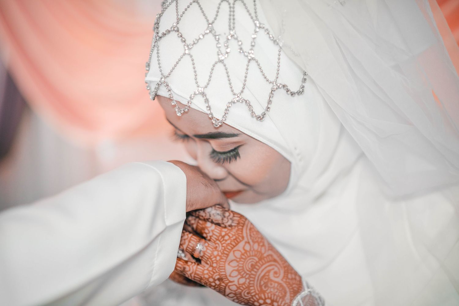 Bride with detailed henna and embellished white hijab in soft romantic lighting.