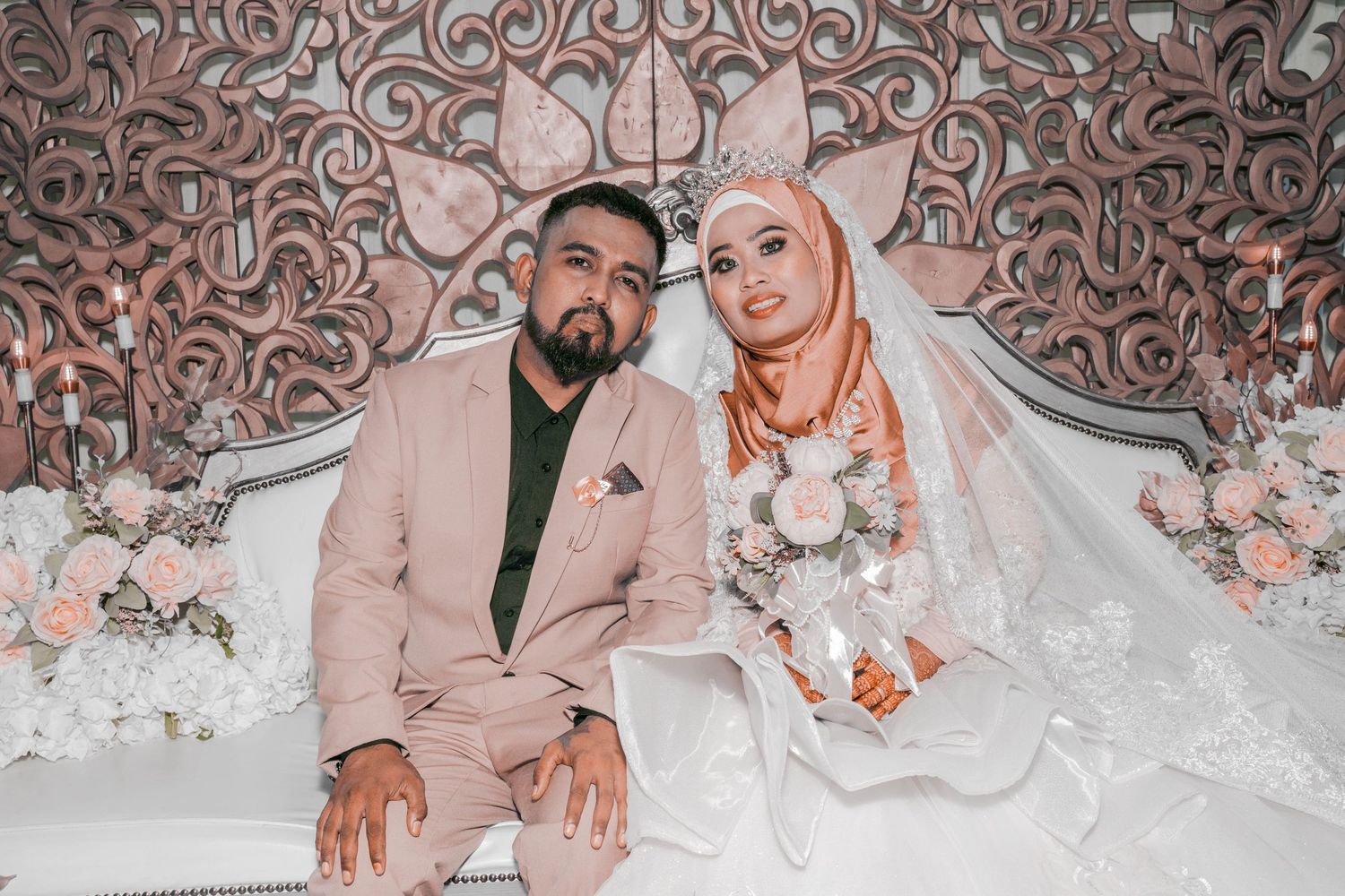Newlyweds seated together against ornate rose-gold patterned backdrop at wedding reception.