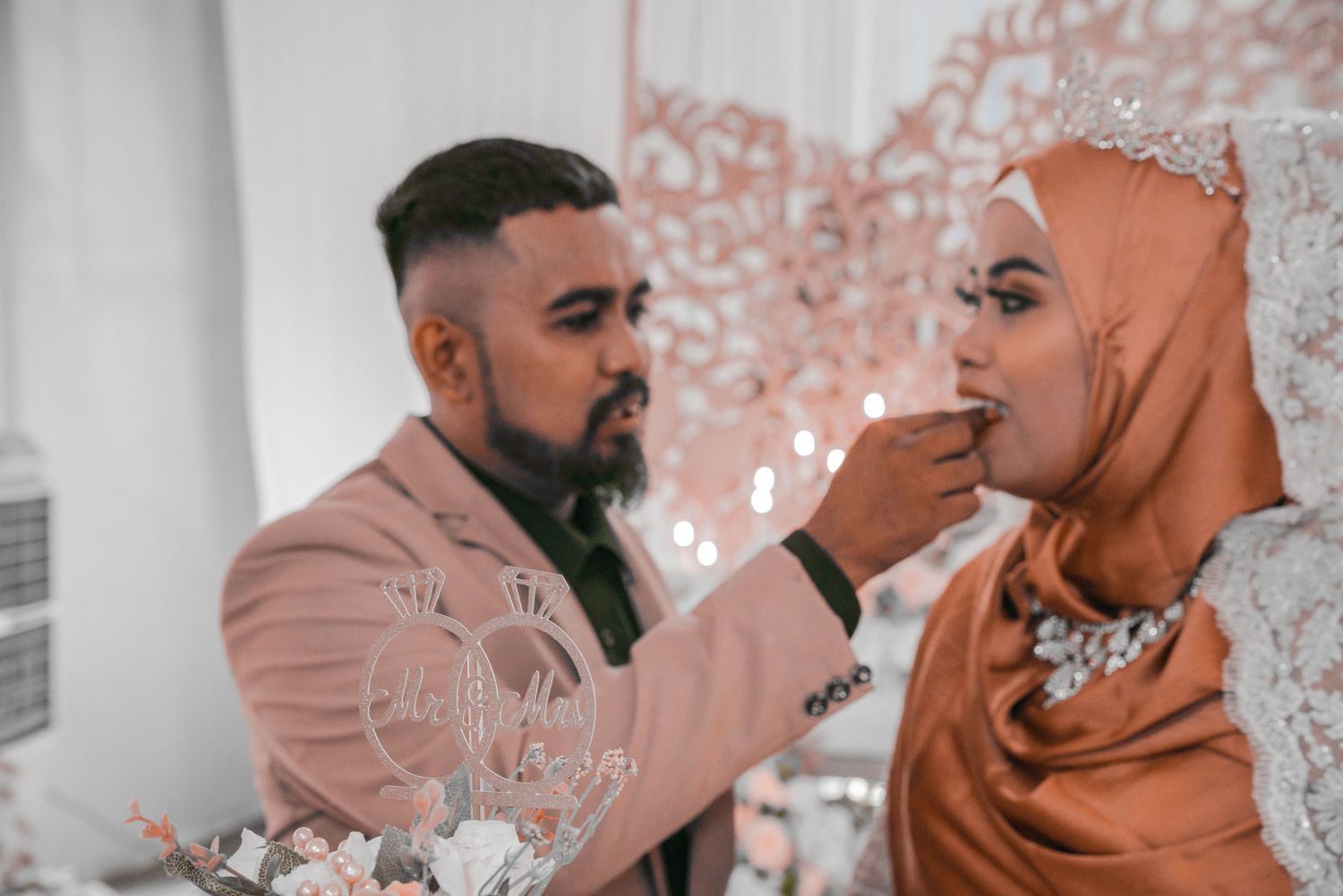 Couple sharing cake during wedding reception against decorative rose-gold backdrop.