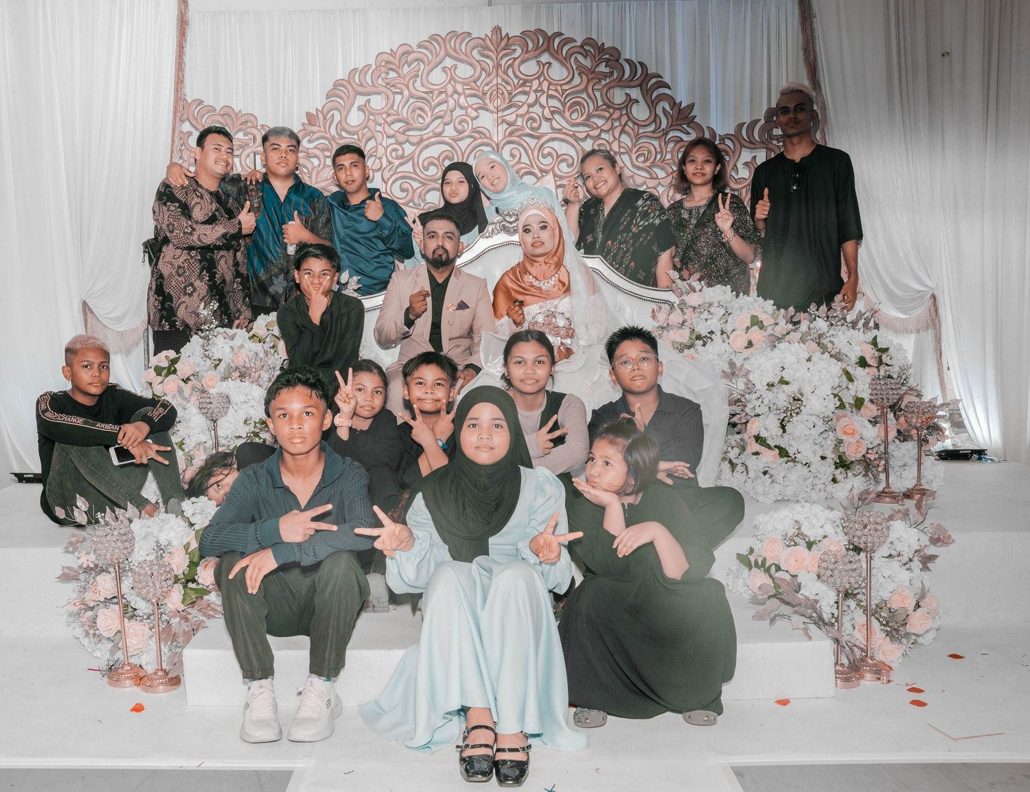Wedding party group photo with guests arranged on decorated white floral backdrop.