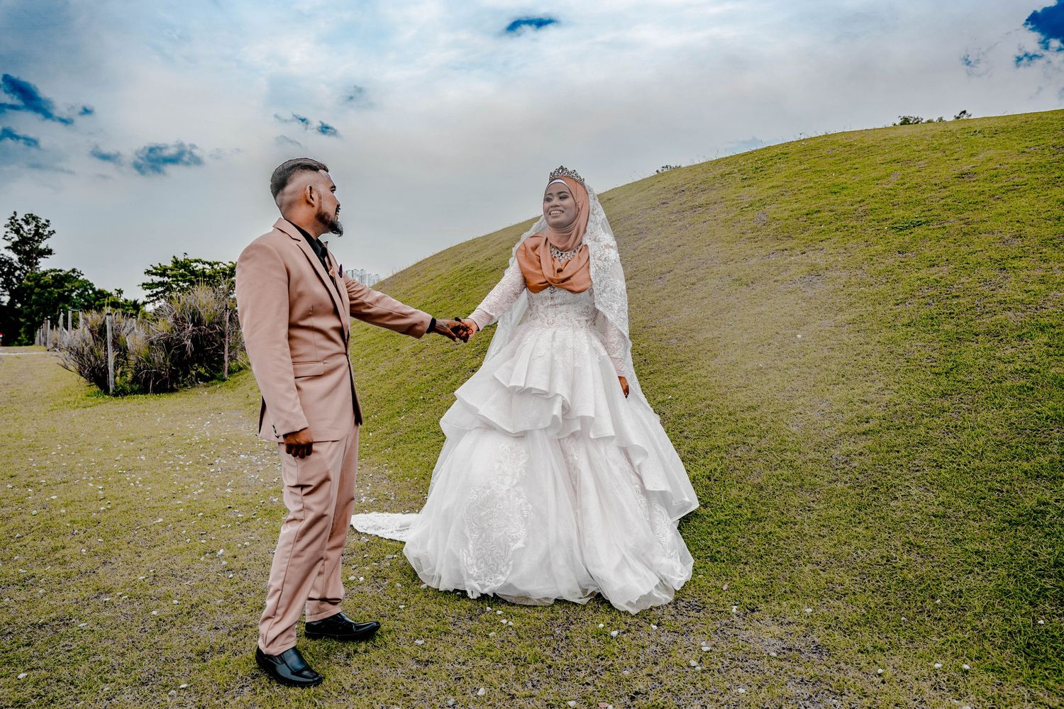 Couple in formal attire posing on green hillside during outdoor wedding photoshoot.