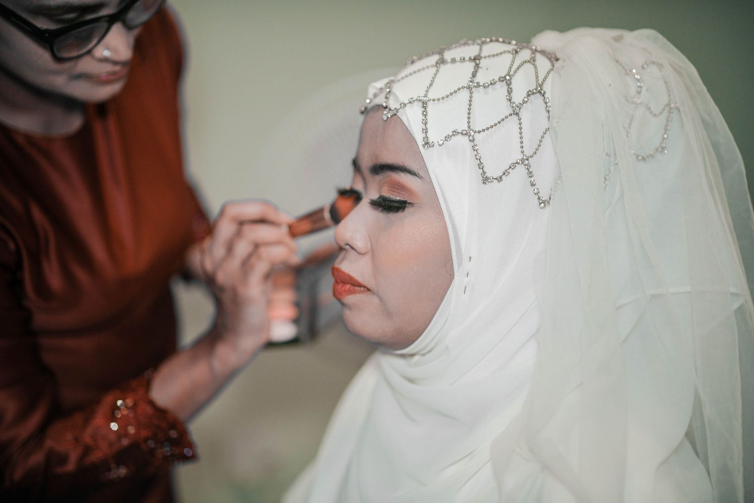 Wedding preparation showing makeup application with white hijab and veil.