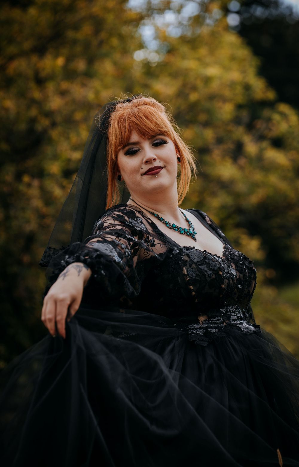 Dark romantic bridal portrait in black lace gown against autumn foliage background.
