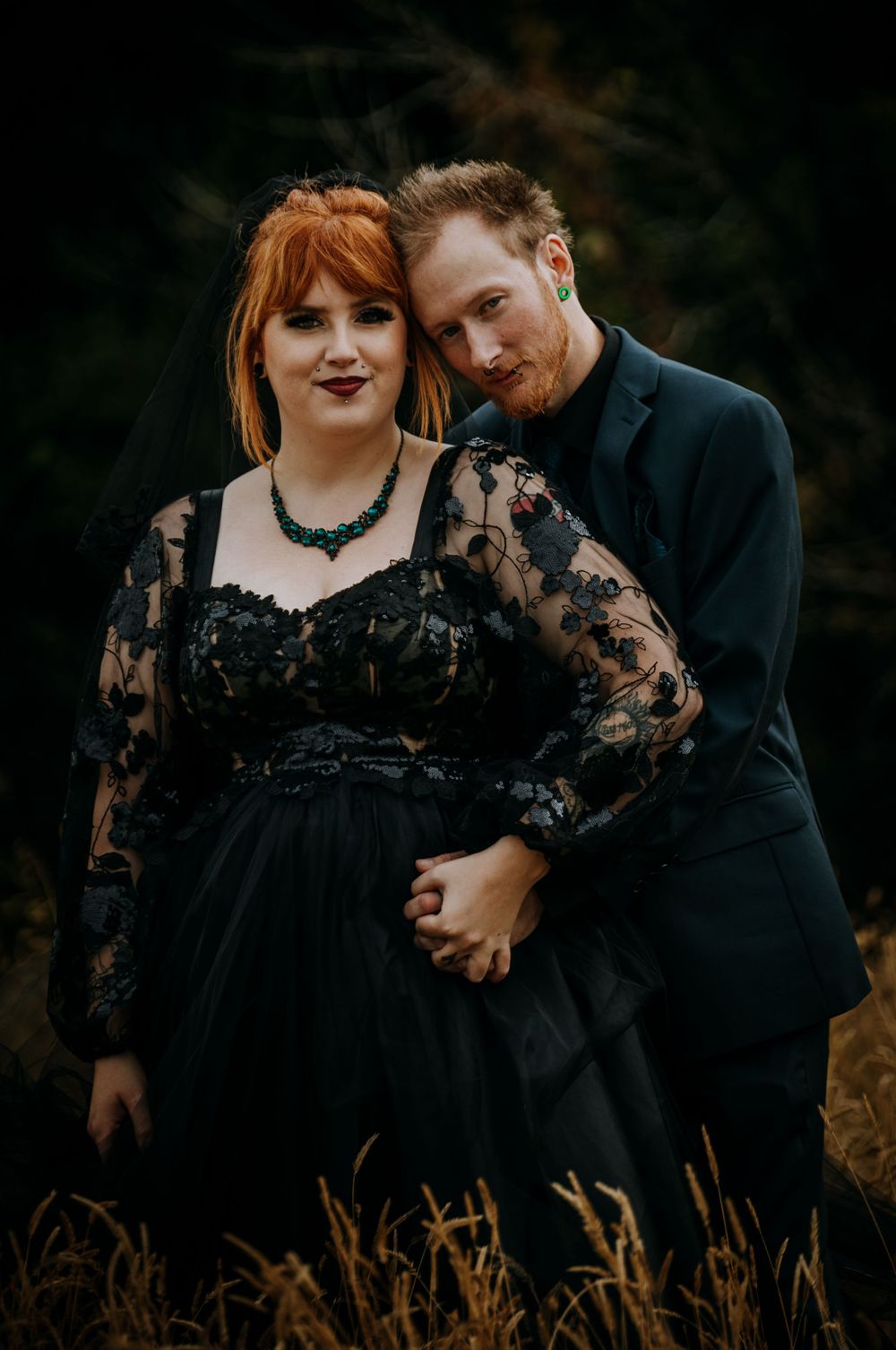 Couple in gothic wedding attire pose together in outdoor autumn setting.