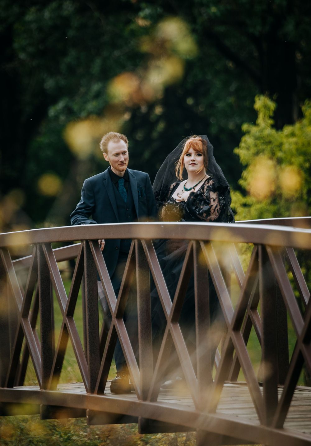 A couple shares a romantic moment while standing on a wooden bridge in a park setting.