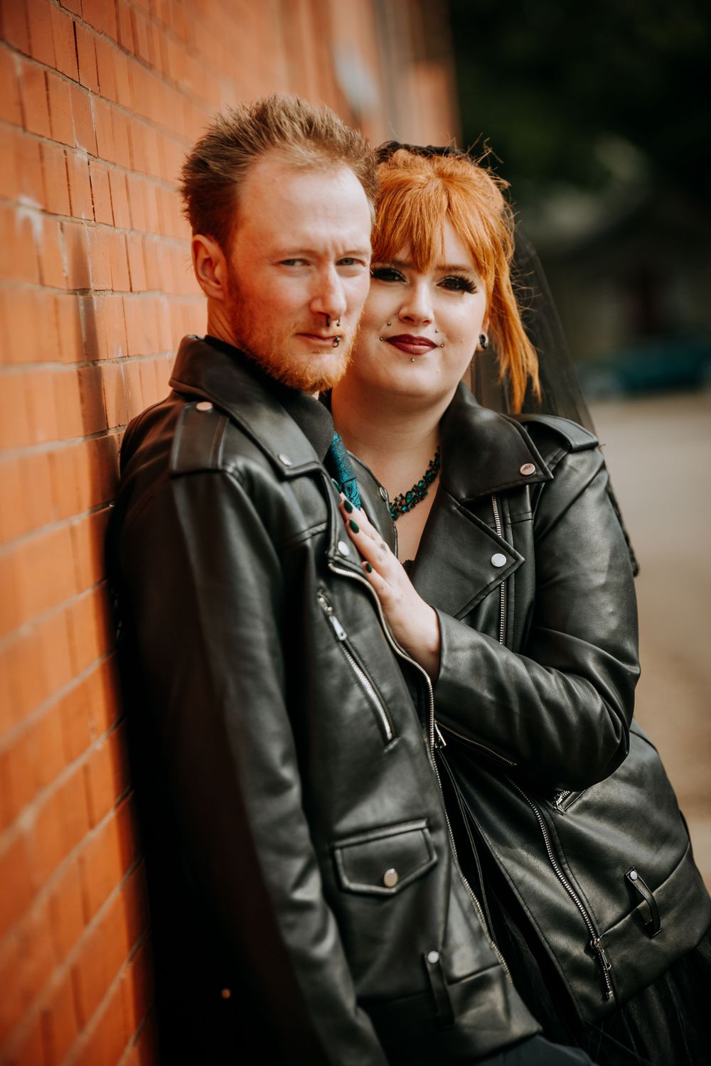 A couple in matching leather jackets poses intimately against a brick wall background.