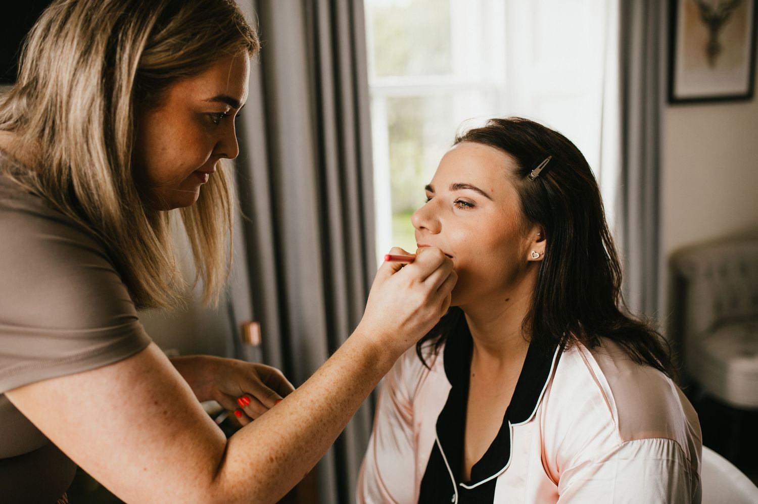 Makeup artist applies lipstick to client during professional beauty session in natural lighting.