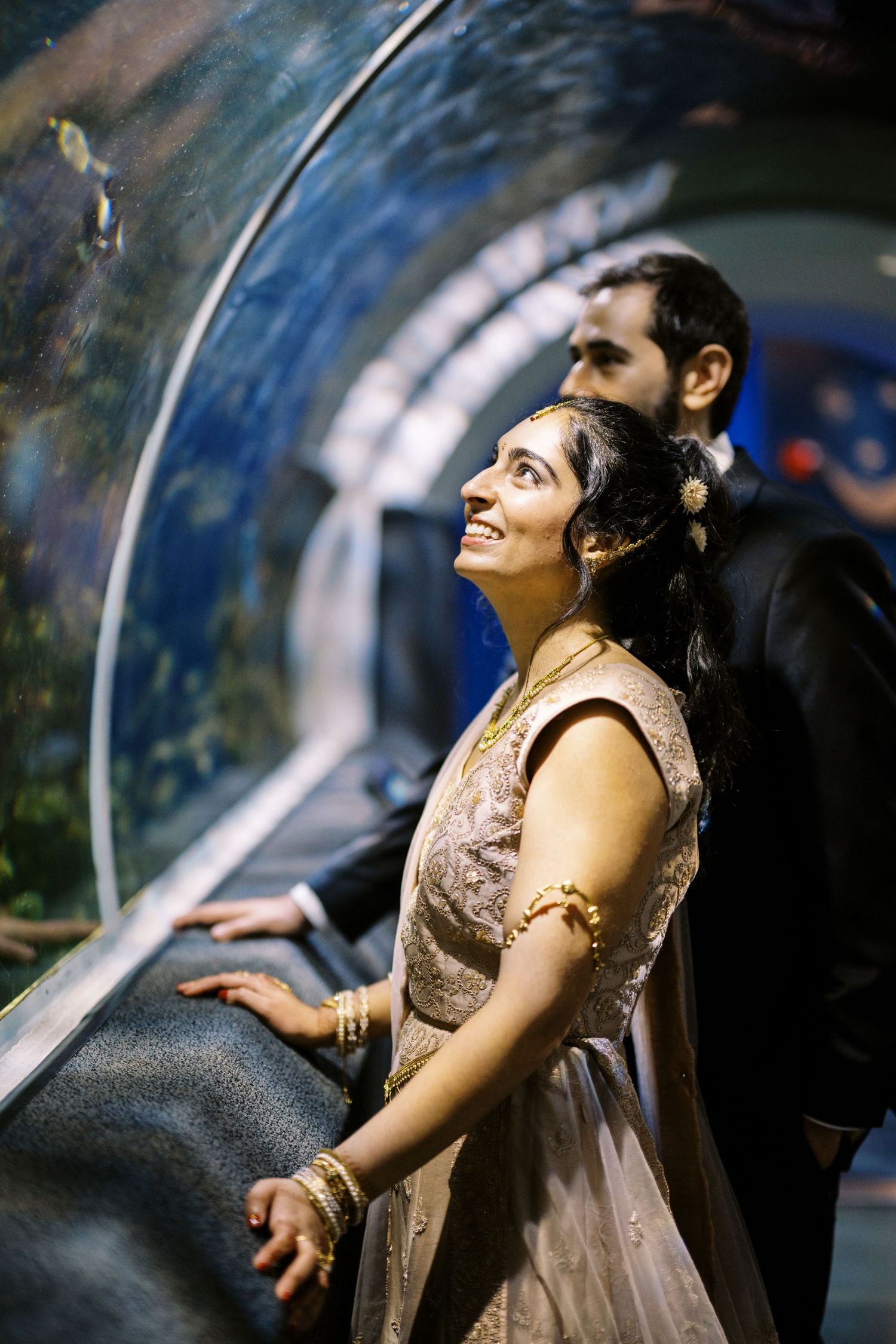 A couple admires marine life in an underwater tunnel at an aquarium, with the woman wearing an elegant beige gown.