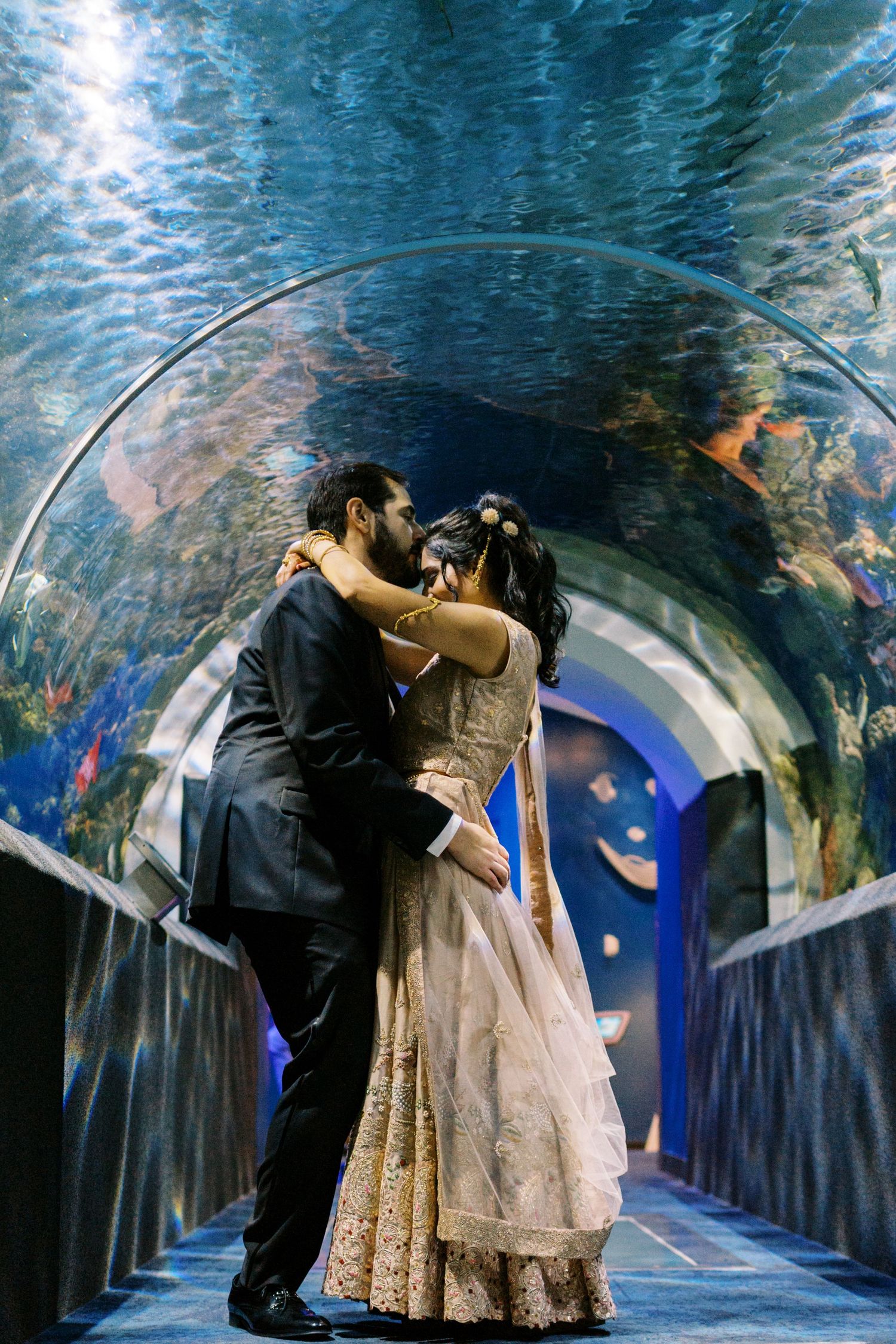 A romantic moment shared between a couple in formal attire under a blue aquarium tunnel surrounded by fish.