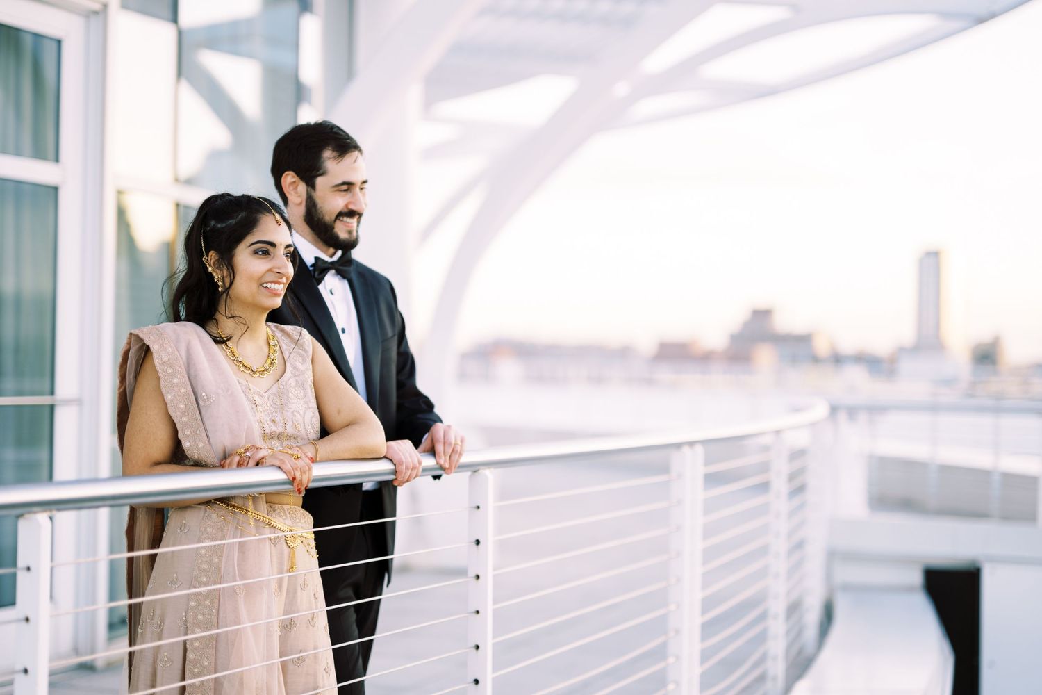 A couple in formal attire shares a tender moment on a balcony overlooking a city skyline during golden hour.