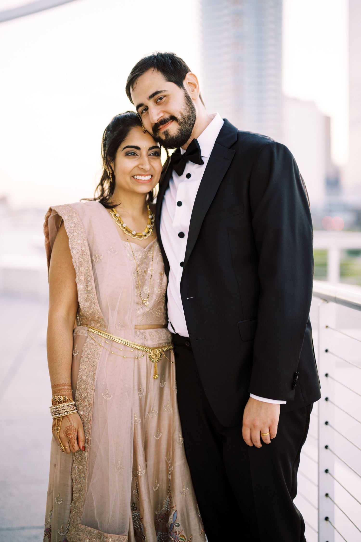 A formal couple poses together against a bright background, with her in a beige dress and him in a tuxedo.