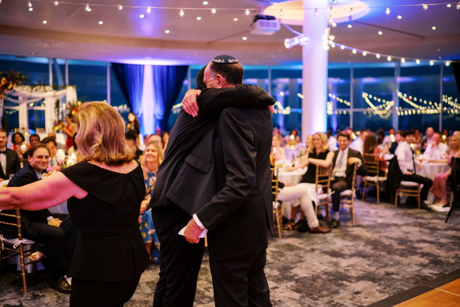 Couples dancing together at an elegant indoor reception with string lights and guests seated in the background.
