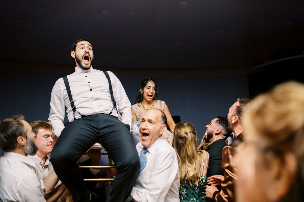 Wedding guests celebrate on a crowded dance floor with people lifted up on shoulders during an energetic party moment.