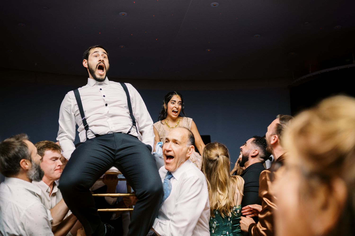 Wedding guests celebrate on a crowded dance floor with people lifted up on shoulders during an energetic party moment.