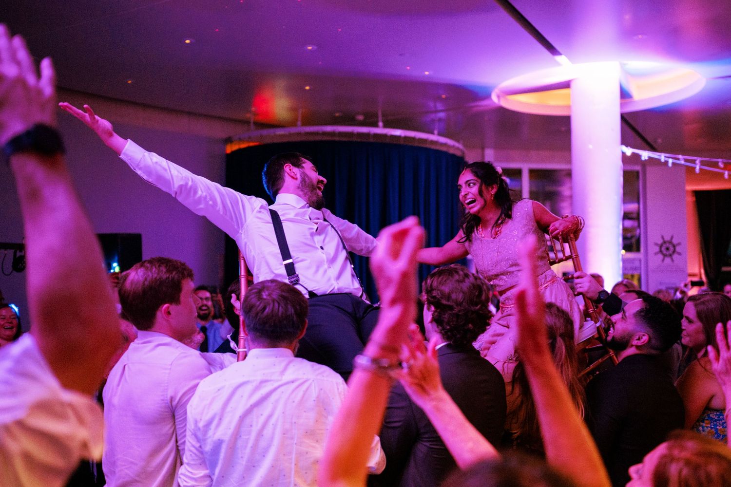A crowd dances under purple lighting at a festive celebration with people being lifted up during the party.