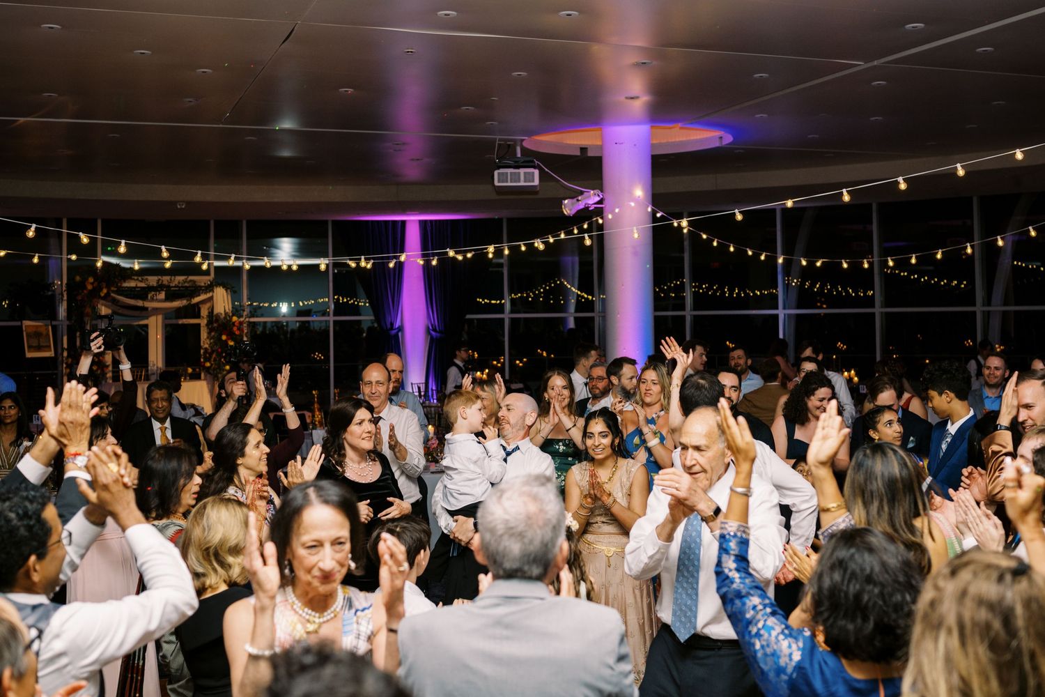 Wedding guests dance and celebrate under purple lighting at an indoor reception venue.