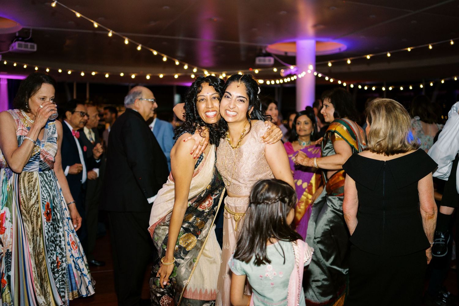 A festive wedding reception with guests dancing under purple lighting and string lights.