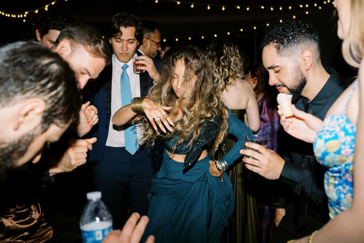 People dancing and celebrating at a nighttime party under string lights at an outdoor event.