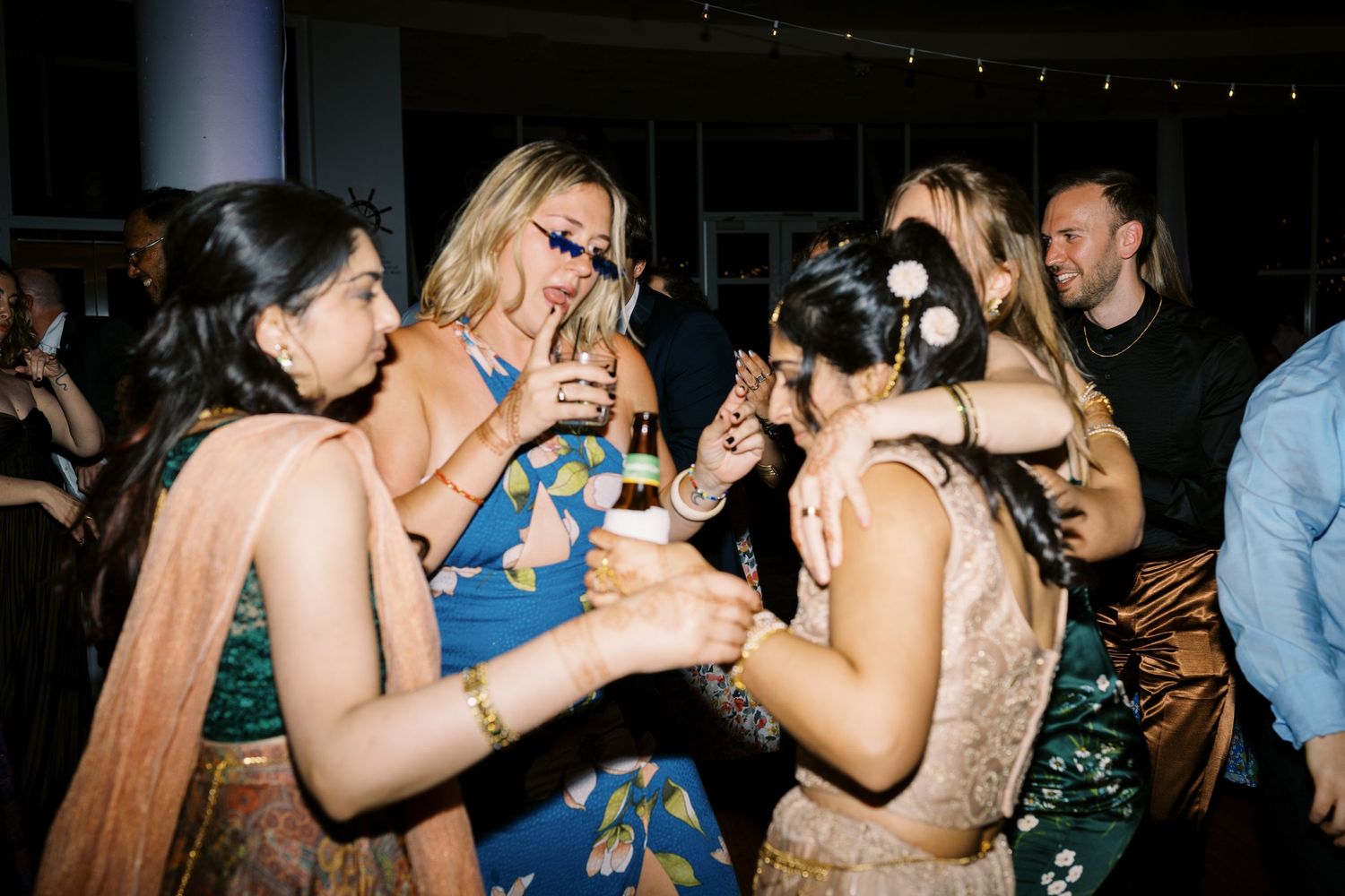 Party guests celebrating and dancing together with drinks in hand at a dimly lit nighttime event.