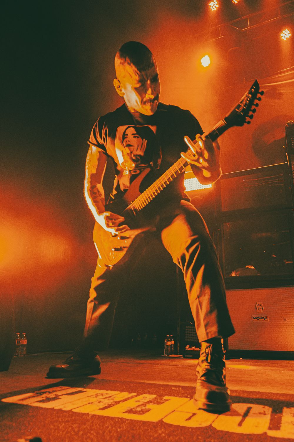 Musician performs on stage in dramatic orange lighting while playing electric guitar during live concert performance.