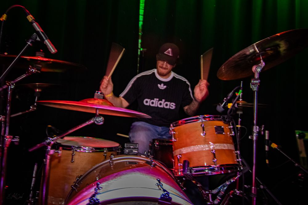 Drum kit setup on stage illuminated by theatrical red and green spotlights during a live performance.