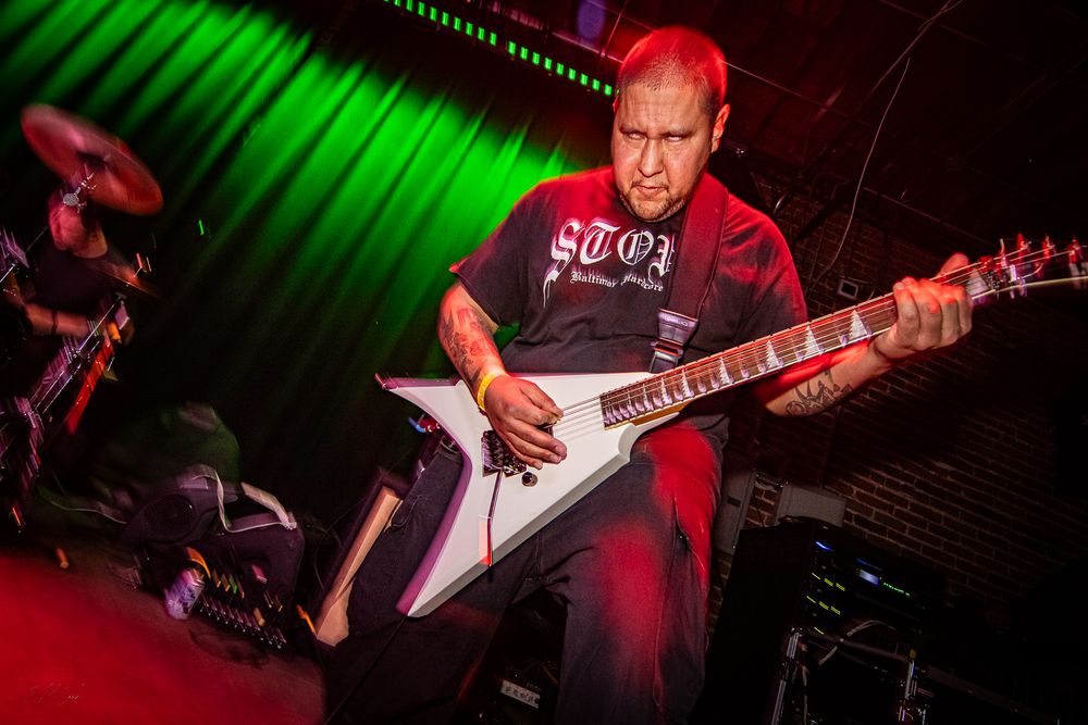 Guitarist plays a white Flying V electric guitar on stage with dramatic red and green lighting.