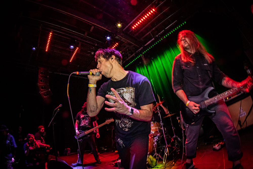Musicians perform on stage at a rock concert with colorful red and green lighting effects.