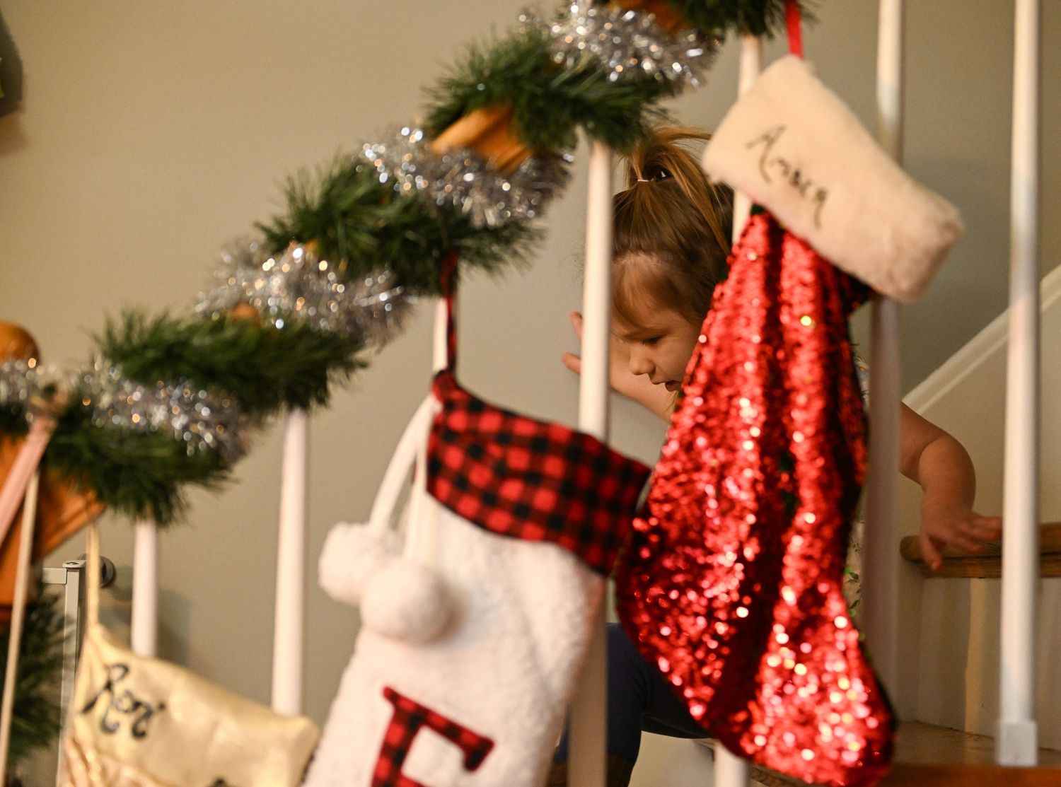A little girl climbs down the stairs behind Christmas stockings and garland hanging off the railing