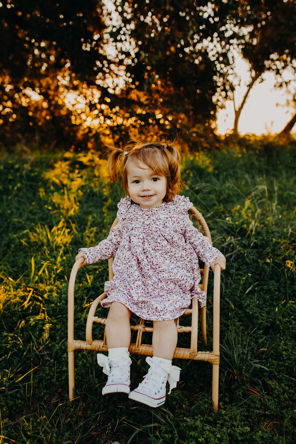 A small figure seated on a wooden chair at sunset wearing a floral dress and white shoes.
