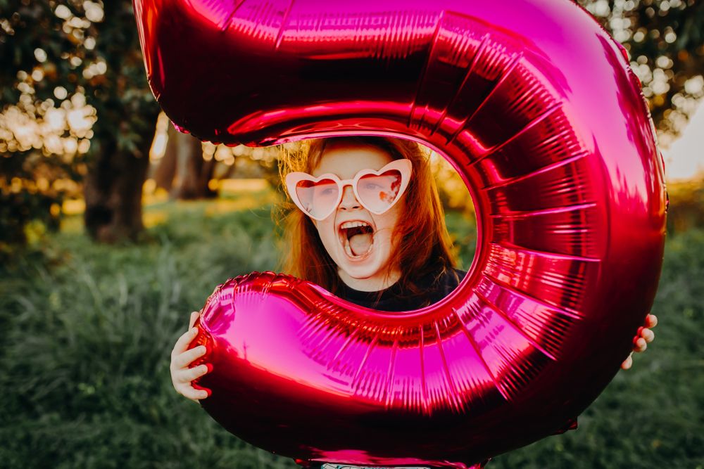 Person expressing excitement while holding large pink number 5 balloon outdoors.