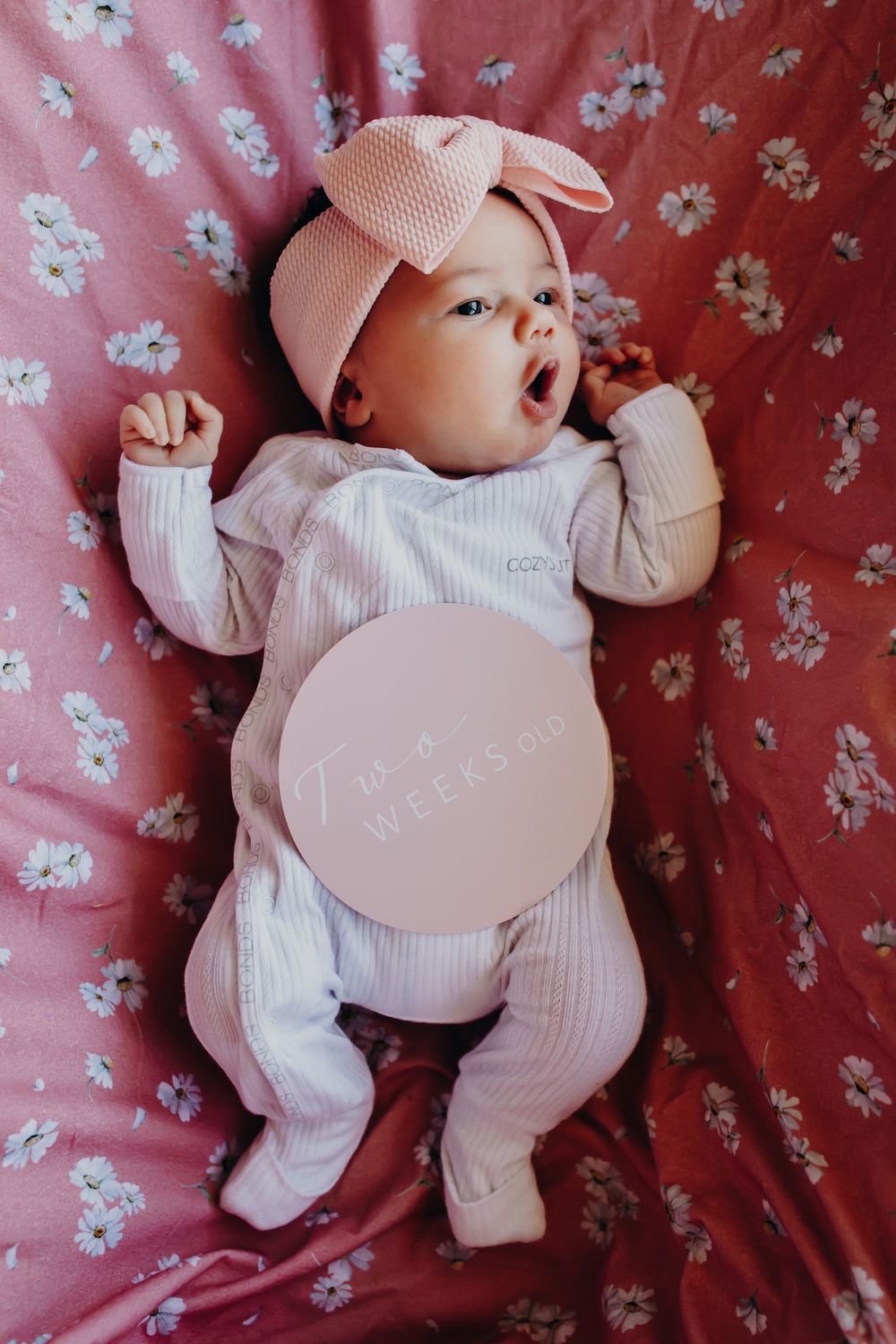 Baby wearing pink bow headband lies on floral background in white onesie.