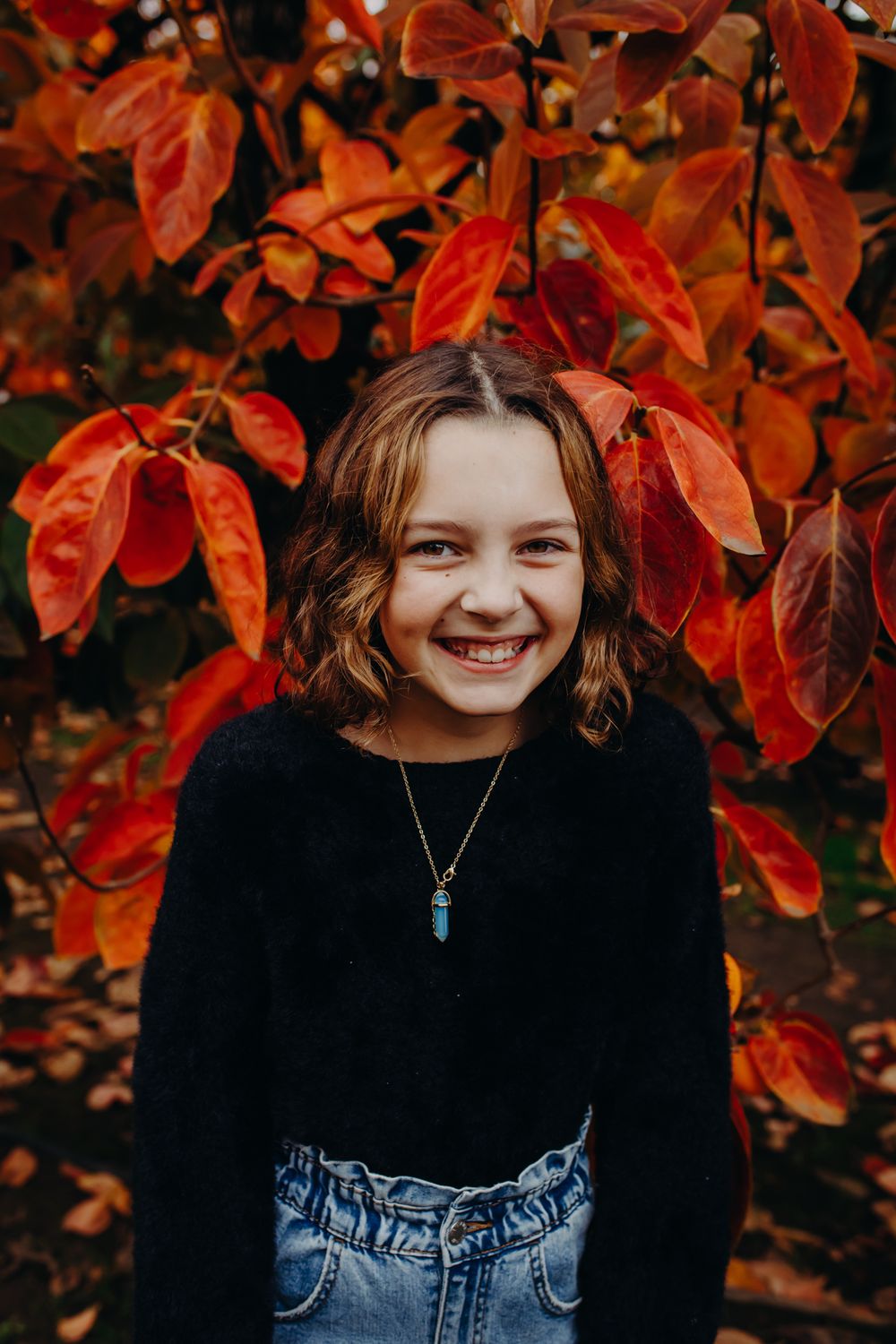 Portrait against vibrant red fall foliage wearing a black sweater and pendant necklace.