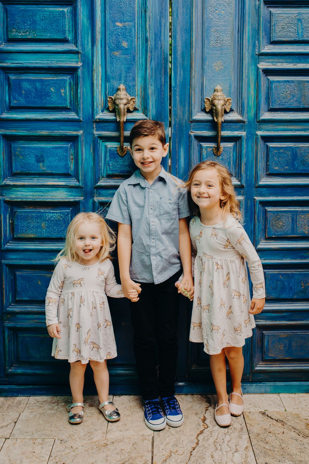 Three children stand together holding hands in front of ornate blue vintage doors.