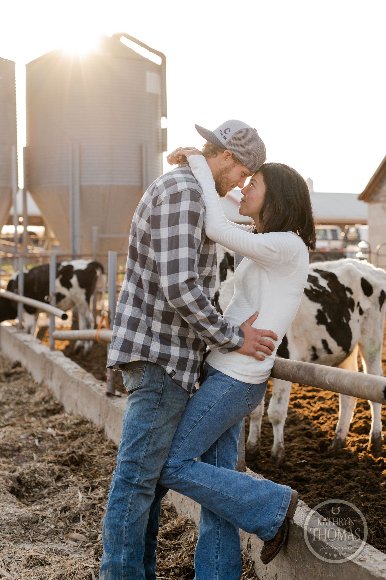 A loving embrace between a couple on a dairy farm with cows and silos in the background.