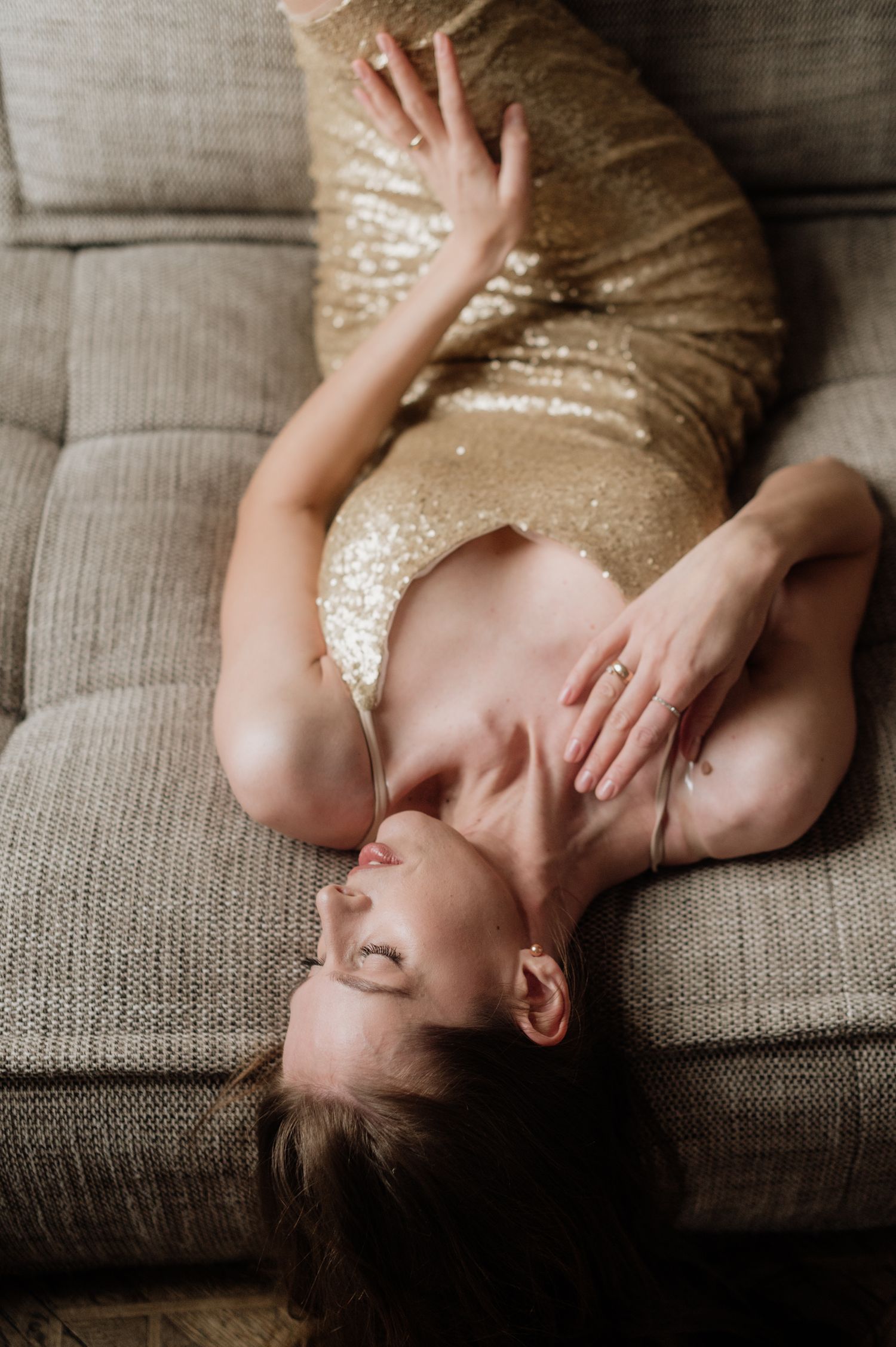 Artistic boudoir portrait of someone reclining on a grey couch in a sparkly gold dress.