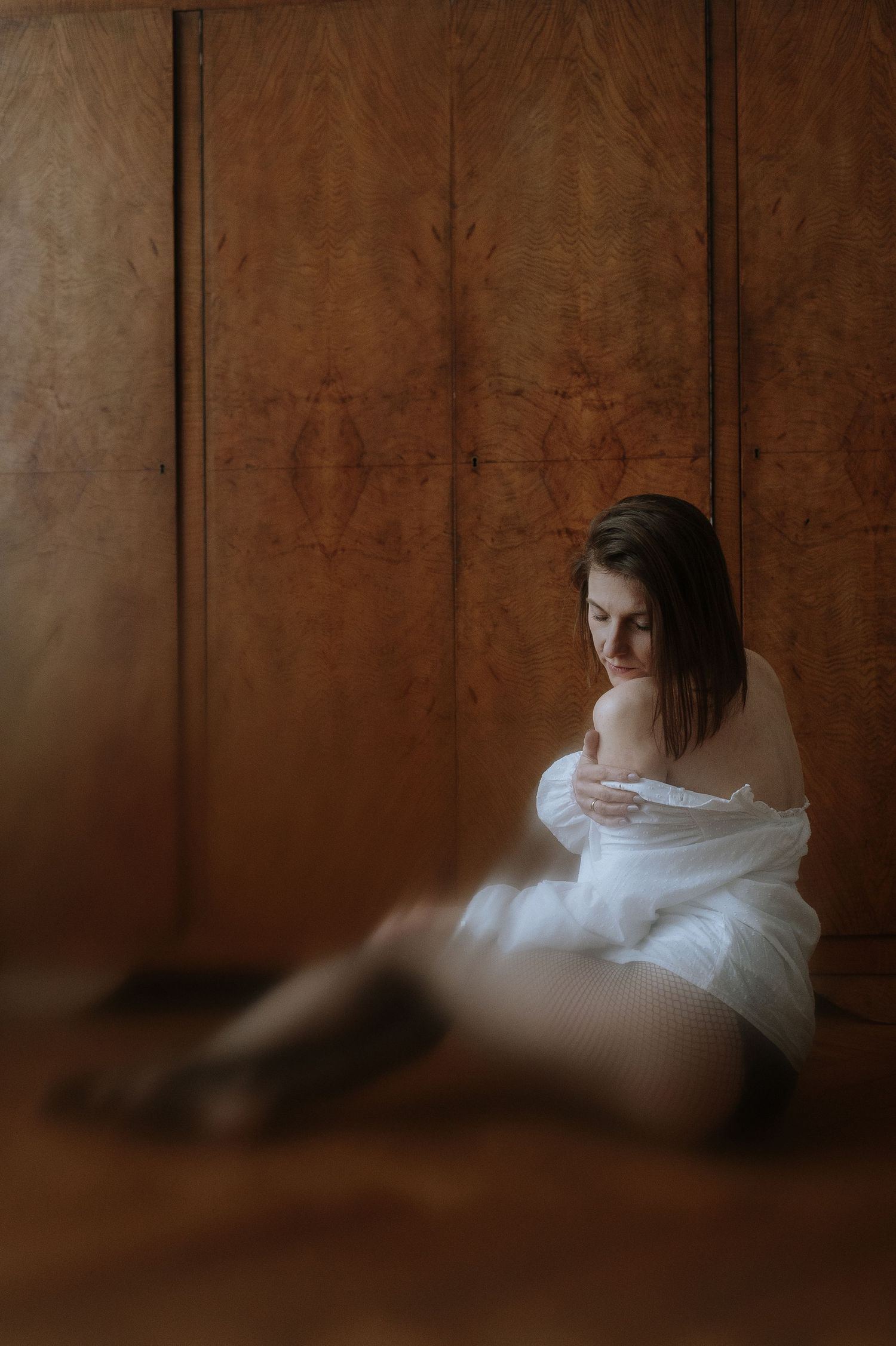 Figure in white fabric seated against dark wooden paneled wall in contemplative pose.