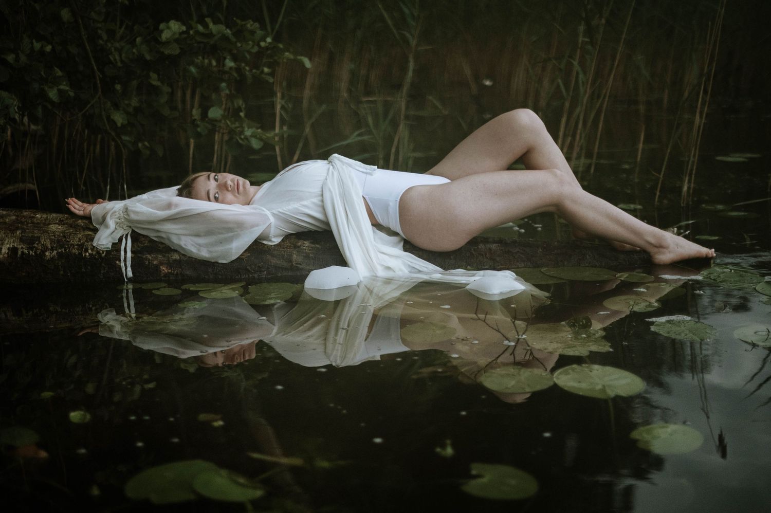Ethereal figure lying gracefully near water with lily pads in moody lighting.