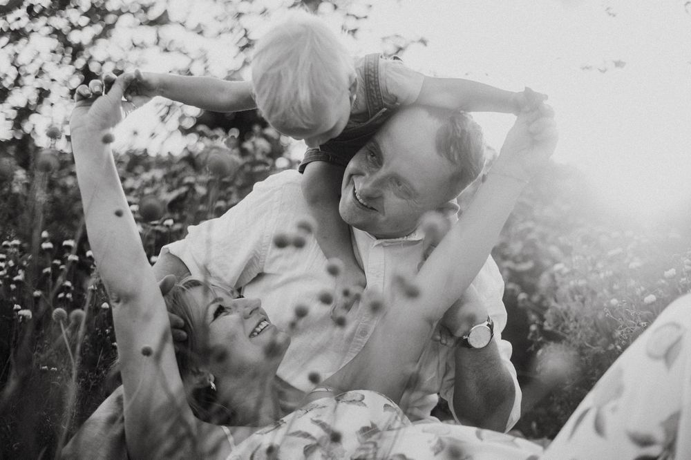 Black and white photo of a joyful family playing outdoors in a sunlit field, parents laughing with young child.