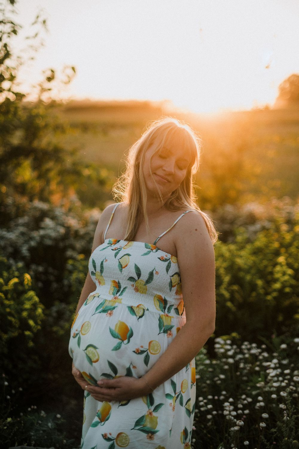 Pregnant woman in floral sundress cradling baby bump at golden hour sunset in a lush green field.