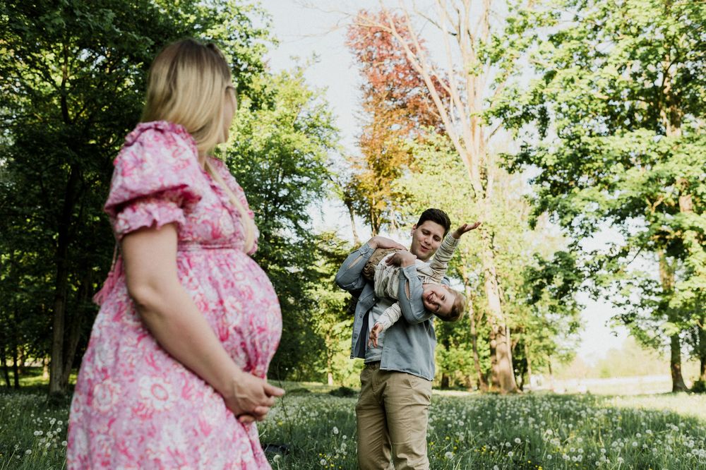 Pregnant woman in pink floral dress stands in park while man holds baby near lush green trees.