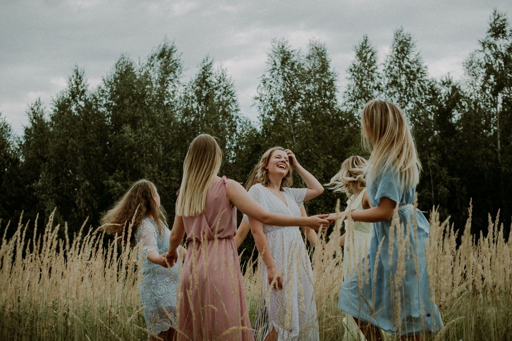 A group in light blue and white dresses dances together joyfully in a golden wheat field at sunset.
