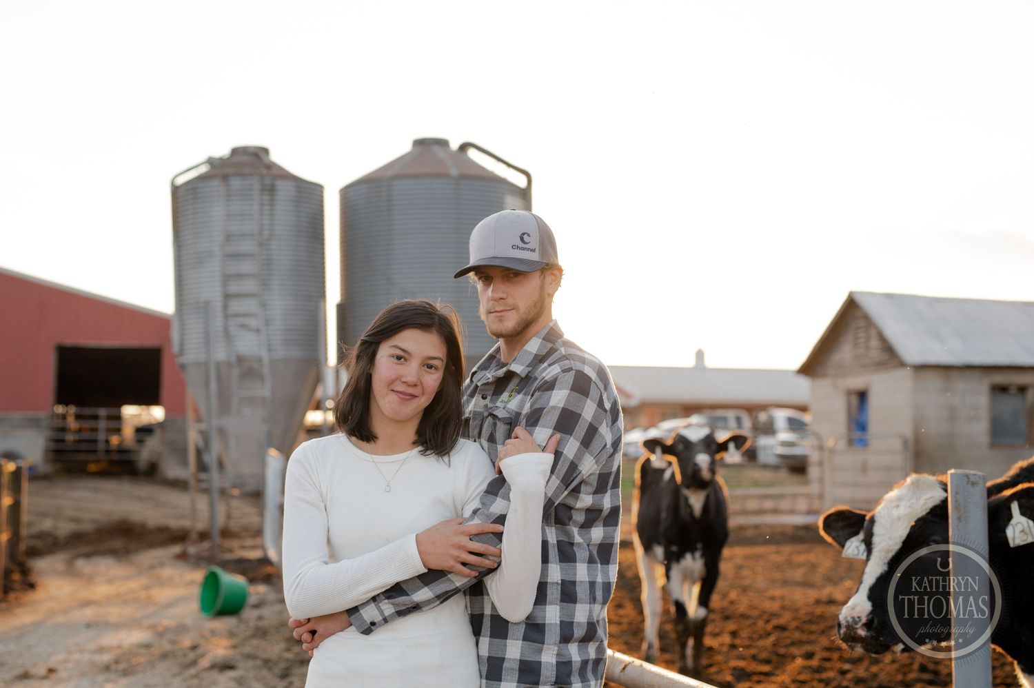 A couple poses near grain silos on a dairy farm with cattle visible in the background.