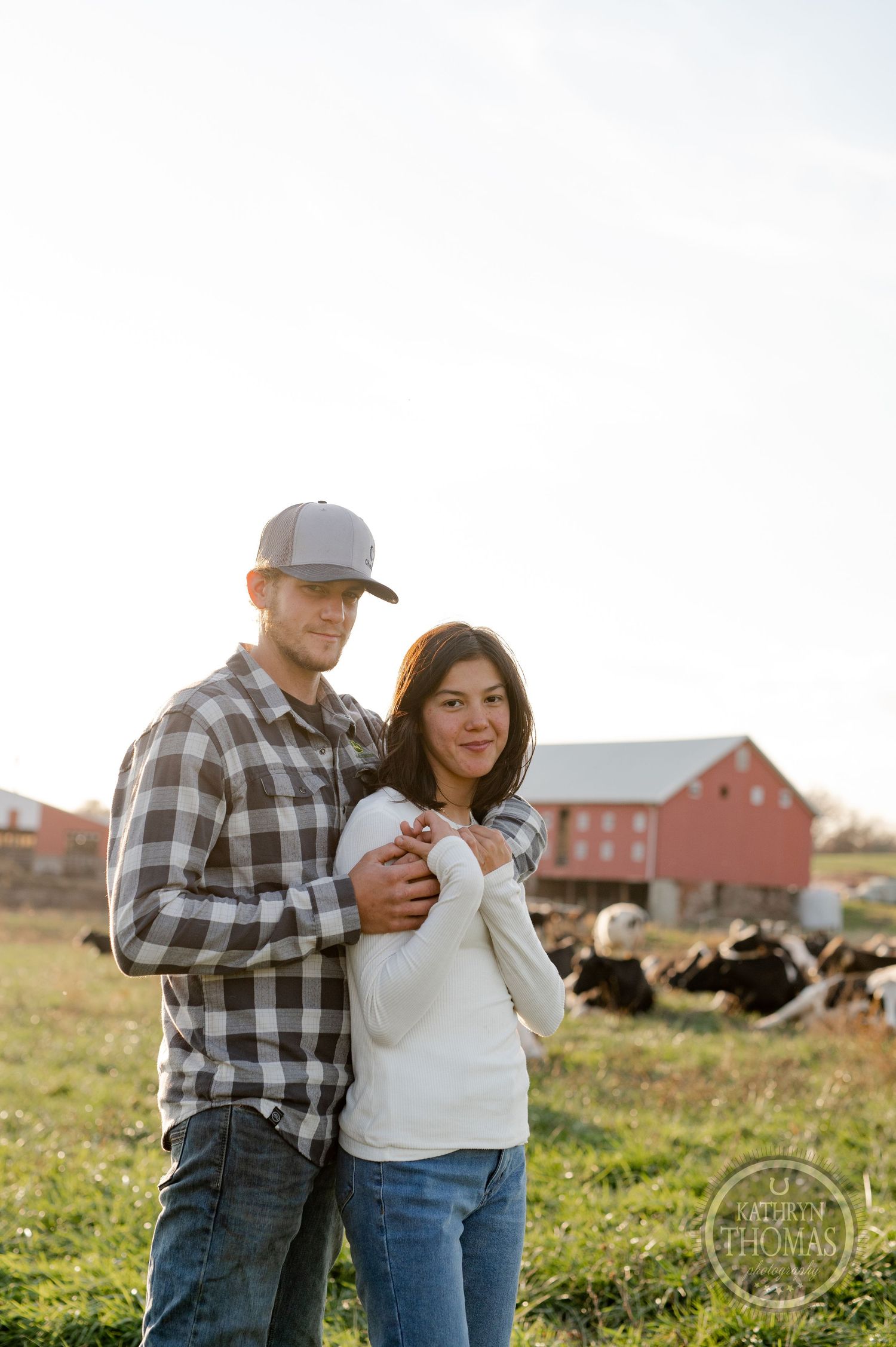 A couple embraces on a rural farm with a red barn and grazing cattle in the background during golden hour.